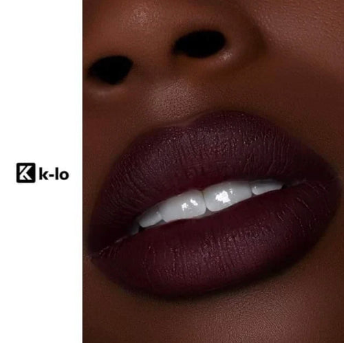 Close-up of a person's lips wearing a deep matte burgundy lip gloss, with part of the nose and teeth visible. The left side has a white vertical band with the visible brand text 'k-Lo'.
